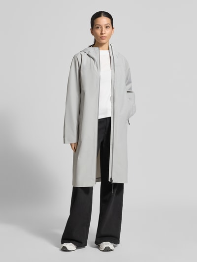 Frauenmodel trägt Mantel mit Zweiwege-Reißverschluss und Kapuze von PEAK PERFORMANCE in Hellgrau, glatte Struktur, Relaxed Fit, Ganzkörperansicht von vorne.