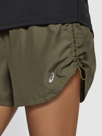 Frauenmodel trägt olivgrüne Shorts mit elastischem Bund, reflektierendem Logo und Reißverschlusstasche von ASICS TIGER, Nahaufnahme Oberschenkel, Modell 'NAGINO', gerade Passform, seitliche Eingrifftaschen, strukturierte Textur, Kontext: Damen Shorts, Bundweite 34 cm, Innenbeinlänge 14 cm bei Größe S.