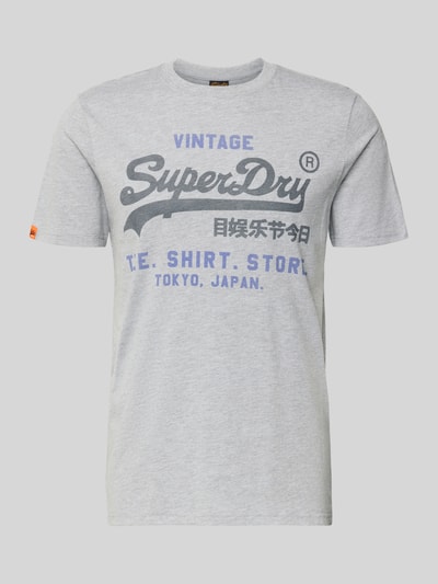 Mężczyznamodel nosi Relaxed Fit T-Shirt mit Logo-Stitching Modell 'Heritage' od Superdry z bawełnianej mieszanki, jasnoszary, nadruk z przodu, okrągły dekolt, długość tyłu w rozmiarze M: 71 cm, Ganzkörperansicht von vorne.