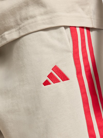 Männermodel trägt Regular Fit Sweatshorts aus Baumwoll-Mix von adidas Sportswear, beige mit roten Kontraststreifen, Logo-Stitching, Nahaufnahme Oberkörper.