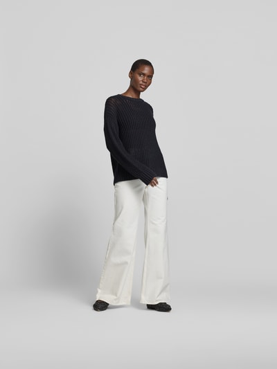 Frauenmodel trägt Pullover in Strick-Optik von Juvia in dunklem Blau, kombiniert mit weißer Hose, semitransparent, Ganzkörperansicht von vorne.