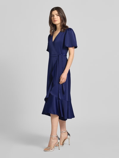 Vrouwenmodel draagt Cocktailjurk met volantzoom model 'THEA' van YAS, satijn, getailleerde fit, wikkel-look met strik, V-hals, vlindermouwen, donkerblauw, midi-lengte, detailopname van voren.