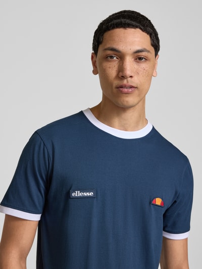Mężczyzna nosi T-Shirt mit Logo- und Label-Patch Modell 'RINGETTO' od Ellesse, czysta bawełna, krój regular fit, okrągły dekolt, granatowy z białym wykończeniem, długość pleców w rozmiarze M: 72 cm, zbliżenie na górną część ciała, przód.