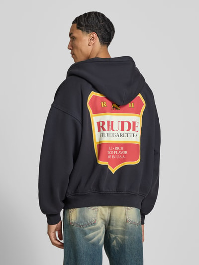 Männermodel trägt Oversized Sweatjacke aus katoen van RHUDE, zwart met opvallende labelprint, kangoeroezak, tweewegritssluiting, Ruglengte maat M 64 cm, Schouderbreedte 45 cm, Mouwlengte 56 cm, Ganzkörperansicht Rückansicht.