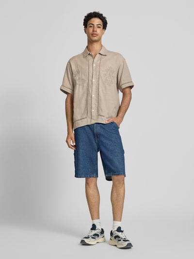 Männermodel trägt Relaxed Fit Freizeithemd aus Lyocell-Mix Modell 'Menton' von Jack & Jones in Beige mit floralem Loch-Stitching, Kurzarm, Kentkragen, durchgehender Knopfleiste, kombiniert mit Jeansshorts; Ganzkörperansicht von vorne.