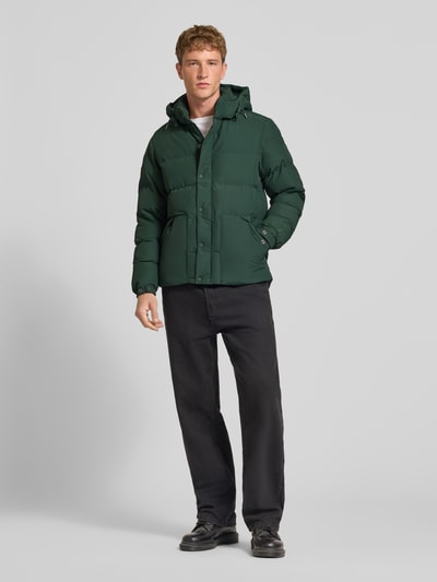 Männermodel trägt dunkelgrüne Jacke mit Kapuze Modell 'CLASSIC EVEREST' von Superdry, gerade Passform, Ganzkörperansicht von vorne.