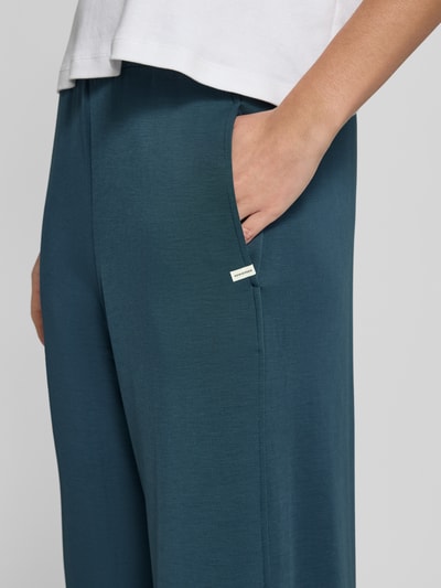 Frauenmodel trägt türkisfarbene Sweatpants aus Modal-Mix mit Label-Detail von SCHIESSER, sichtbar elastischer Bund und Eingrifftaschen, Nahaufnahme Oberkörper.