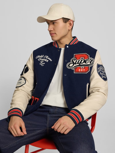 Męski model nosi College-Jacke mit Patches und Eingrifftaschen od Superdry, mieszanka bawełny, krój regular fit, stójka, listwa guzikowa, wpuszczane kieszenie, naszywki, widok całego ciała z przodu. Długość tyłu w rozmiarze M: 65 cm, szerokość ramion w rozmiarze M: 45 cm, szerokość w rozmiarze M: 49 cm, długość rękawów w rozmiarze M: 70 cm.