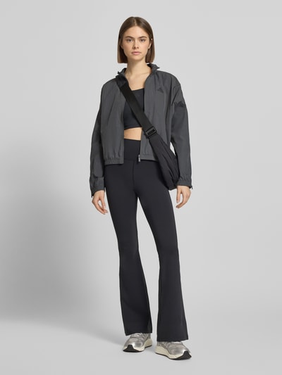 Modelka nosi Regular Fit Jacke mit Stehkragen von adidas Sportswear, ciemnoszara, logo-print, zamki, elastyczne zakończenia, długość tyłu w rozmiarze S: 54 cm, szerokość ramion w rozmiarze S: 64 cm, szerokość klatki piersiowej w rozmiarze S: 64 cm, długość rękawa w rozmiarze S: 48 cm, widok całej sylwetki z przodu.