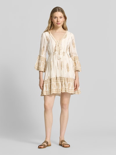 Frauenmodel trägt Minikleid mit Allover-Paisley-Print von Apricot aus Viskose-Mix, Taillierte Passform, V-Ausschnitt, 3/4-Arm, Schnürverschluss, Rüschenbesatz, Rückenlänge bei Größe S 83 cm, Brustweite 33 cm, Ärmellänge 49 cm, helle und beigefarbene Töne, fließender Stoff, Paisley-Muster, Ganzkörperansicht von vorne.