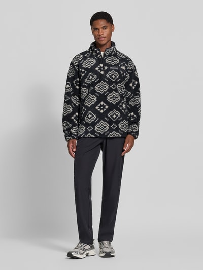 Męski model nosi Regular Fit Pullover mit Logo-Patch od Columbia z polaru, krój regular fit, naszywka z logo, kołnierz zapinany, długość tyłu w rozmiarze M 70 cm, długość rękawów w rozmiarze M 68 cm, wzór geometryczny w odcieniach czerni i bieli, tekstura miękka, całopostaciowy widok z przodu.