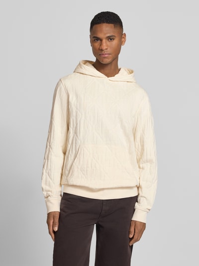Männermodel trägt Hoodie mit Känguru-Tasche und Kapuze von Guess aus reiner Baumwolle im Loose-Fit, helle Farbe, Ganzkörperansicht von vorne.