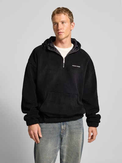 Männermodel trägt Fleecepullover mit Reißverschluss Modell 'Olten' von Pegador in Schwarz mit Kapuze und Känguru-Tasche, Nahaufnahme Oberkörper.