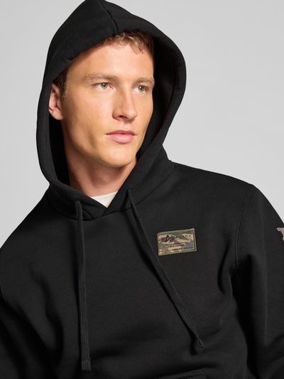 Männermodel draagt Hoodie met labelstitching model 'Camo Sleeve' van Alpha Industries in zwart katoenmix, regular fit, nahaufnahme bovenlichaam.