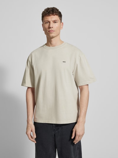Męski model nosi T-Shirt mit Logo-Print Modell 'Gary' od Wood Wood, kremowa bawełna, prosty krój, nadruk z logo, widok z przodu na całą sylwetkę.