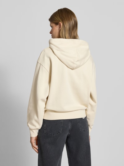 Kobiece modelka nosi bluzę rozpinaną Sweatjacke mit Känguru-Tasche od Carhartt Work In Progress, bawełna, krój relaxed fit, kaptur, wyhaftowane logo, zamek błyskawiczny, kieszeń kangurka, widok całej sylwetki od tyłu.
