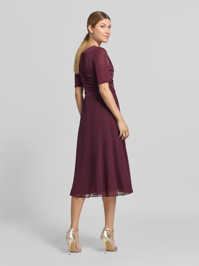 Männermodel trägt Fit and Flare Cocktailkleid mit Wasserfallausschnitt von Vera Mont, sukienka koktajlowa w kolorze bordowym, lekki materiał, gładka faktura, Ganzkörperansicht Rückansicht.