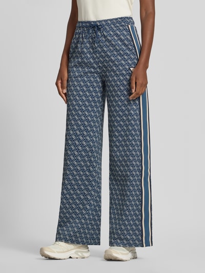 Frauenmodel trägt Sweatpants mit Logo-Muster im Allover-Look von GUESS Activewear in Blau, Wide Leg, elastischer Bund, Nahaufnahme Unterkörper.