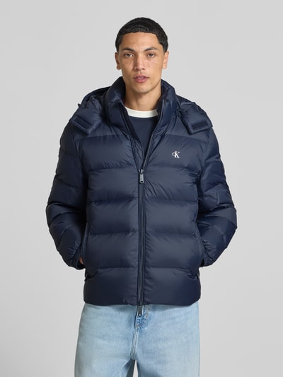 Männermodel trägt Regular Fit Steppjacke mit abnehmbarer Kapuze von Calvin Klein Jeans in Dunkelblau mit matter Oberfläche, Ganzkörperansicht von vorne.