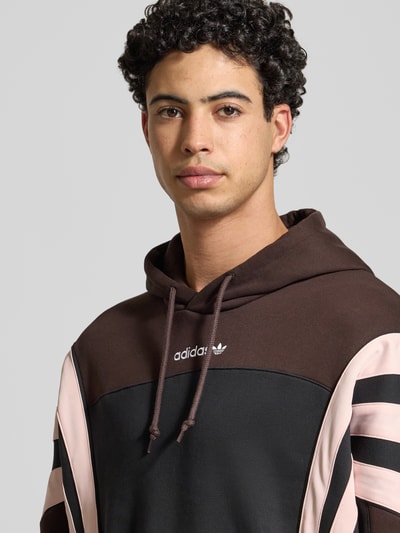 Männermodel trägt Hoodie mit Kapuze von adidas Originals in Braun und Schwarz mit Label-Stitching, Nahaufnahme Oberkörper. Kontext: Herren Hoodie, Lockere Passform/Schnitt:, Kapuze, Tunnelzug, Label-Stitching, Känguru-Tasche, Gerippte-Abschlüsse, Rückenlänge 71 cm, Ärmellänge 66 cm.
