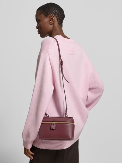 Frauenmodel trägt Crossbody Bag aus echtem Leder von Victoria Beckham in Bordeaux mit Rundum-Reißverschluss, Label-Print in metallic, Verstellbarer Schulterriemen, Dornschließe, Hauptfach, Breite 21 cm, Länge 13 cm, Tiefe 7 cm, Ganzkörperansicht Rückansicht.