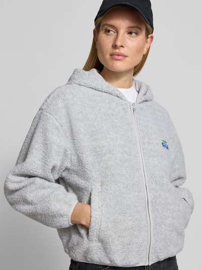 Frauenmodel trägt Jacke aus Teddyfell Modell 'HOKTOWN' von American Vintage in Hellgrau mit Kapuze, Reißverschluss, Eingrifftaschen, Label-Detail, Ganzkörperansicht von vorne.