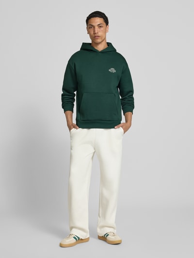 Männermodel trägt Hoodie mit Logo-Print und Känguru-Tasche von REVIEW aus Baumwoll-Mix, Relaxed-Fit, Kapuze, gerippte Abschlüsse, Ganzkörperansicht von vorne.