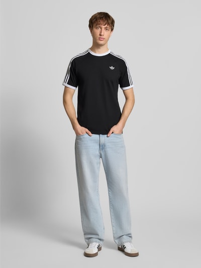 Männermodel trägt T-Shirt mit Label-Stitching von adidas Originals, schwarze Reine Baumwolle, Regular-Fit, Rundhalsausschnitt, Ganzkörperansicht von vorne.
