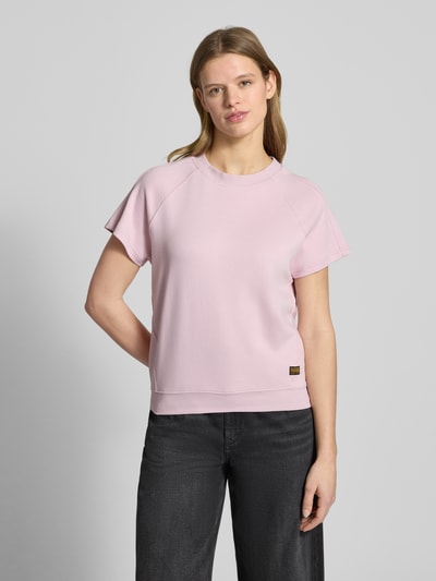 Kobiece modelka nosi Regular Fit T-Shirt mit Logo-Patch Modell 'Raglan' von G-Star Raw z bawełny, okrągły dekolt, krój regular fit, naszywka z logo, elastyczne wykończenie, długość tyłu 57 cm w rozmiarze S, jasnoróżowy kolor, gładka faktura, widok całej sylwetki z przodu.