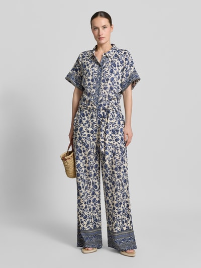 Vrouwenmodel draagt dames jumpsuit van LOLLYS LAUNDRY, zuiver viscose, relaxed fit, model 'Mathilde', all-over look, overhemdkraag, strikceintuur, ruglengte maat S 46 cm, borstwijdte 44 cm, mouwlengte 17 cm, totale lengte 160 cm, binnenbeenlengte 77 cm, blauw-wit bloemdessin, recht en losvallend, Ganzkörperansicht von voren.