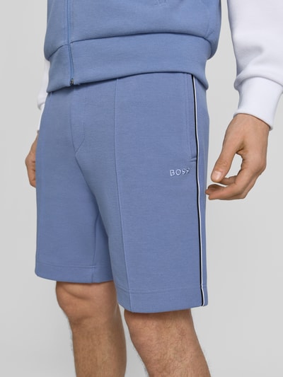 Männermodel trägt Regular Fit Shorts aus Baumwoll-Mix Modell 'T_JOIN Headlo' von BOSS Green in Blau mit Optik: Unifarben, Kontraststreifen, Label-Stitching-Applikation, Bügelfalten, elastischem Bundabschluss, seitlichen Reißverschlusstaschen, Materialverarbeitung: Stretch; Nahaufnahme Oberschenkel.