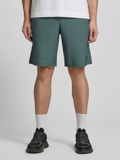 Mężczyzna nosi Regular Fit Shorts mit Strukturmuster von EA7 Emporio Armani w zielonym kolorze, krój regular fit, elastyczny materiał, szerokość w rozmiarze M 41 cm, długość nogawki wewnętrznej 26 cm, widok całej sylwetki z przodu.