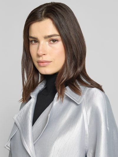 Frauenmodel trägt Jacke aus Viskose-Mix mit Reverskragen und Gürtel von Marc Cain, silber glänzende Textur, Nahaufnahme Oberkörper.