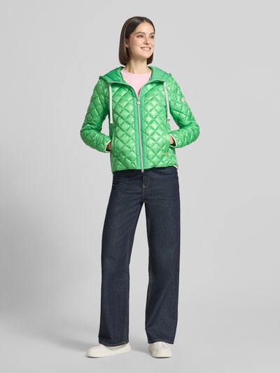Frauenmodel trägt grüne Regular-Fit Jacke in Stepp-Optik Modell 'YOSIE' von Frieda & Freddies mit Kapuze, Tunnelzug, Eingrifftaschen, Reißverschluss, Logo-Applikation, elastischen Ärmelabschlüssen, Ganzkörperansicht von vorne. Rückenlänge 61 cm, Schulterbreite 46 cm, Brustweite 49 cm, Ärmellänge 60 cm bei Größe 36.