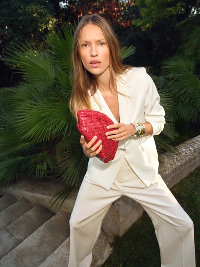 Frauenmodel trägt Regular Fit Blazer mit floralen Applikationen von Christian Berg Woman Selection im Elastischer-Material-Mix, Reverskragen, Knopf, in heller Farbe, kombiniert mit heller Hose, lebendige Rot-Akzente durch Tasche, natürliche Umgebung, Ganzkörperansicht von vorne.