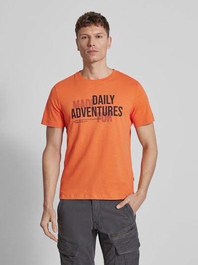 Mężczyznamodel nosi Regular Fit T-Shirt aus reiner Baumwolle von camel active w kolorze pomarańczowym, nadruk z napisem, okrągły dekolt, krój regular fit, długość tyłu w rozmiarze M: 68 cm, męski t-shirt, widok na cały przód.