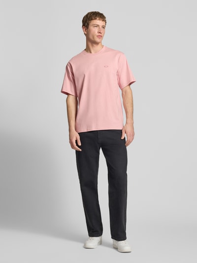 Männermodel trägt T-Shirt mit Label-Detail und Rundhalsausschnitt von Dickies in Rosa aus Baumwolle, Relaxed Fit, Ganzkörperansicht von vorne.