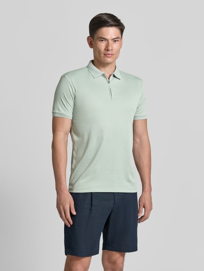 Męski model nosi jasną jednokolorową koszulkę polo Regular Fit model 'FAVE' z mieszanki bawełny ekologicznej marki SELECTED HOMME, krótki rękaw, kołnierzyk polo, krótki zamek błyskawiczny, naszywka z logo, miękka faktura interlock; Ganzkörperansicht von vorne.