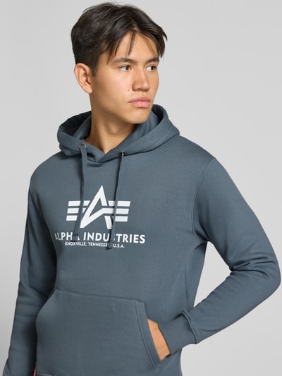 Männermodel trägt Regular Fit Hoodie aus Baumwoll-Mix mit Logo-Print von Alpha Industries, dezentes Grau, Nahaufnahme Oberkörper. Herren Hoodie von Alpha Industries, Label am Saum, Kapuze mit Tunnelzug, langärmliges Design, elastische Abschlüsse.