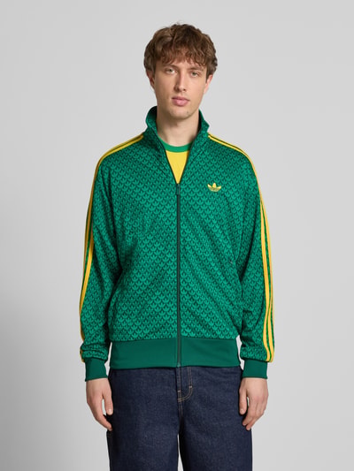 Männermodel trägt Slim Fit Jacke mit Stehkragen Modell 'Firebird' von adidas Originals, grün-gelbes Muster, Logo-Stitching, Ganzkörperansicht von vorne.