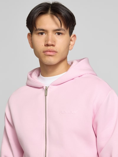 Męski model nosi Sweatjacke mit Känguru-Tasche von REVIEW z mieszanki bawełny, krój relaxed fit, zamek błyskawiczny, kieszeń kangurka, jasnoróżowy, faktura gładka, Nahaufnahme górnej części ciała.