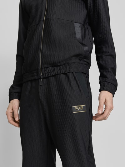 Mężczyzna nosi Regular Fit Sweatpants mit Strukturmuster od EA7 Emporio Armani, czarny, elastyczny materiał, kieszenie, zbliżenie na górną część ciała.