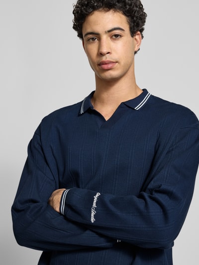 Männermodel trägt Strickpolo aus reine katoen Modell 'JORNIAGARA' van Jack & Jones, donkerblauw, losse fit, V-hals met contrasterende kraag, elastische mouwboorden, labeldetail, ruglengte maat M 64 cm, nahaufnahme bovenlichaam.