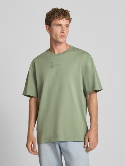 Männermodel trägt T-Shirt mit Logo und Rundhalsausschnitt von Karl Kani in Grün, Relaxed Fit, reine Baumwolle, Ganzkörperansicht von vorne.