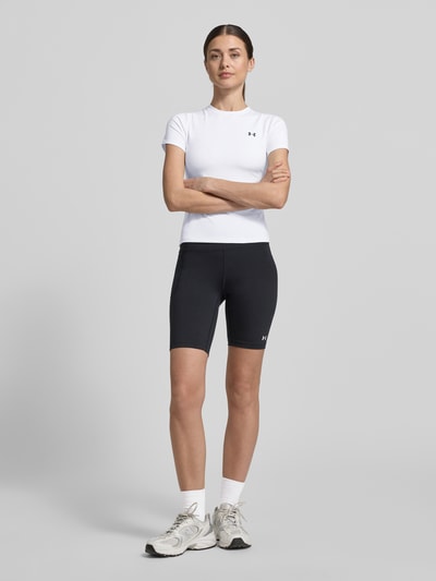 Modelka kobieca nosi T-Shirt mit Rundhalsausschnitt und Logo od Under Armour, biały materiał z domieszką stretchu, krój slim fit, widok całej sylwetki z przodu. Długość pleców w rozmiarze S: 49 cm.