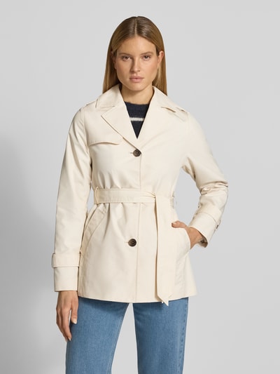 Vrouwenmodel draagt Regular Fit Trenchcoat uit katoenmix van s.Oliver RED LABEL in effen kleuren met labelapplicatie, korte lengte, waterafstotend twill, reverskraag, eenrijige knoopsluiting, tailleriem, zij-ingreepzakken, achterpas, schoudertrensjes, lange mouwen met trensje, binnenzak; kleuren: crème, textuur: glad; geheel van voren.
