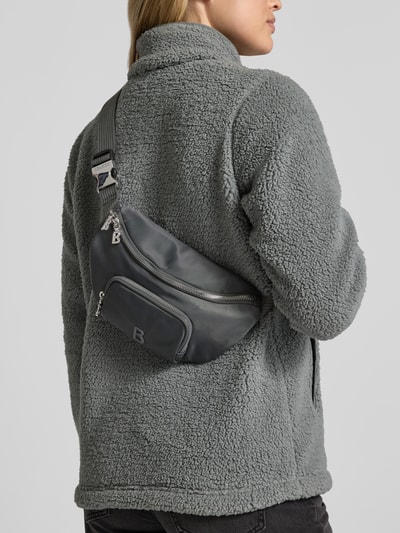Frauenmodel trägt dunkelgraue Bauchtasche mit Label-Detail Modell 'verbier play 1.0 janica' von BOGNER, Detailaufnahme Rückansicht. Hauptfach mit Zweiwege-Reißverschluss, sichtbares Außenfach mit Reißverschluss, strukturierte Oberfläche, breiter Schulterriemen.