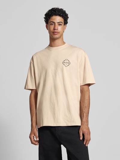Männermodel trägt Oversized T-shirt mit labelprint Modell 'Paide' van Pegador aus puur katoen in beige met zwarte print, ronde hals, oversized fit, ruglengte bij maat M 70 cm, Ganzkörperansicht van voren.