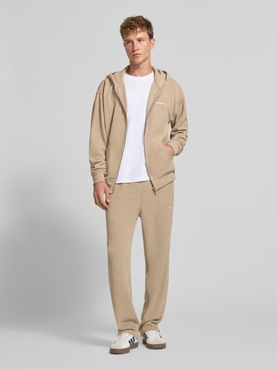 Männermodel trägt Oversized Fit Sweatjacke mit Label-Detail Modell 'Mezzano' von PEGADOR in Beige, weicher Material-Mix, Ganzkörperansicht von vorne. Herren Sweatjacke, getrennte Känguru-Tasche, elastische Abschlüsse, Rückenlänge 70 cm, Ärmellänge 68 cm.