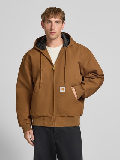 Männermodel trägt Jacke mit Label-Patch und Känguru-Tasche von Carhartt Work In Progress in Braun, Reißverschluss, Kapuze, Nahaufnahme Oberkörper.